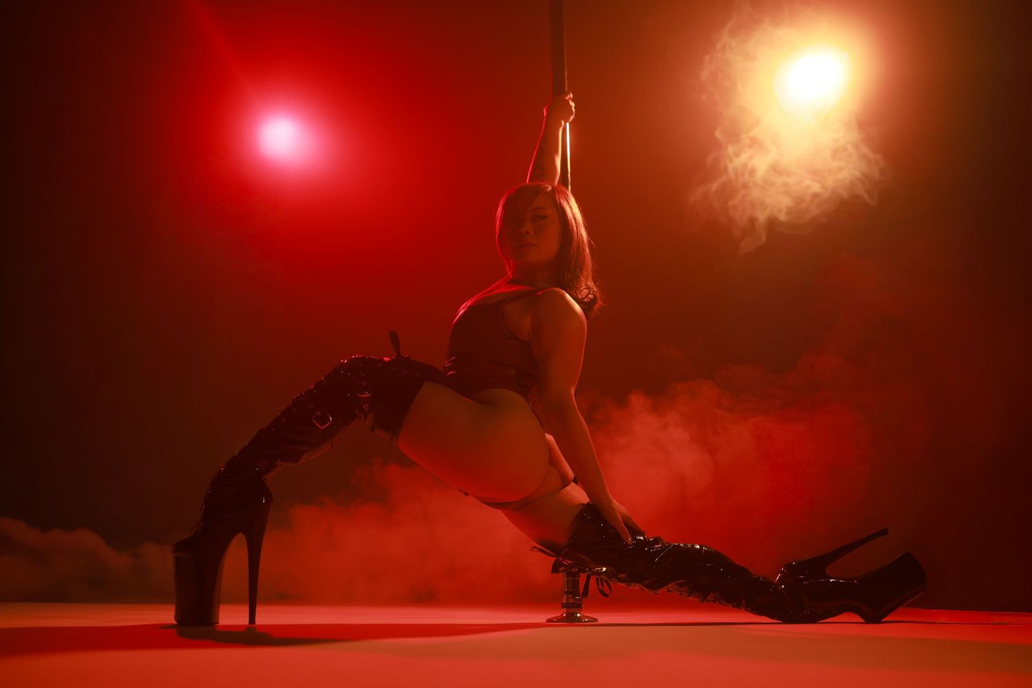A female pole dancer in black boots and lingerie performs an inverted split pose on a pole in red smoky lighting.