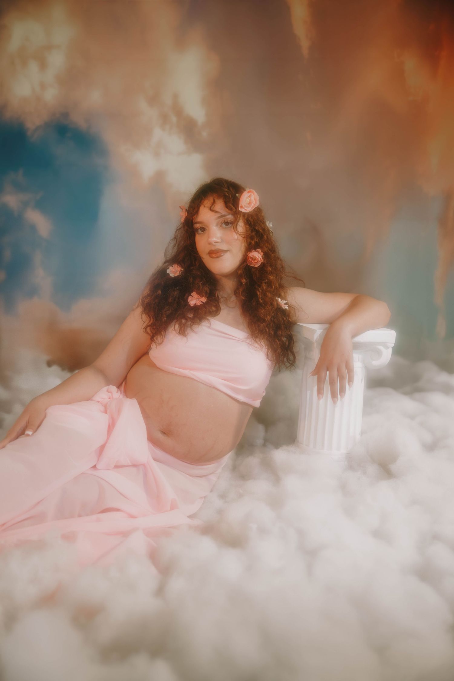 A pregnant woman in pink with flowers in her hair reclines beside a column among soft clouds.