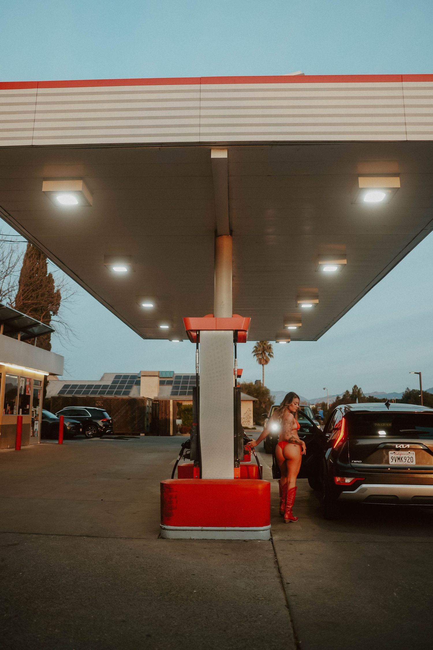 Woman in red outfit stands at illuminated gas station pump at twilight.
