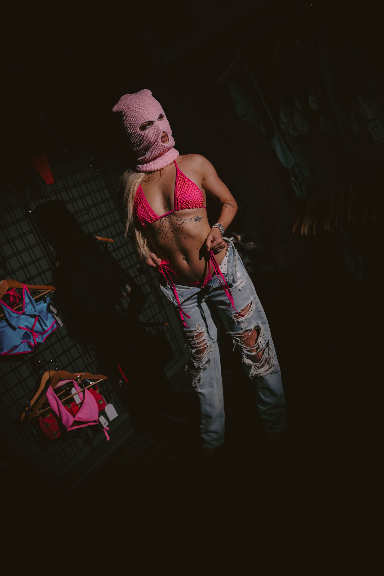 Tattooed model in pink ski mask and bikini top with ripped jeans posing in dark boutique setting.