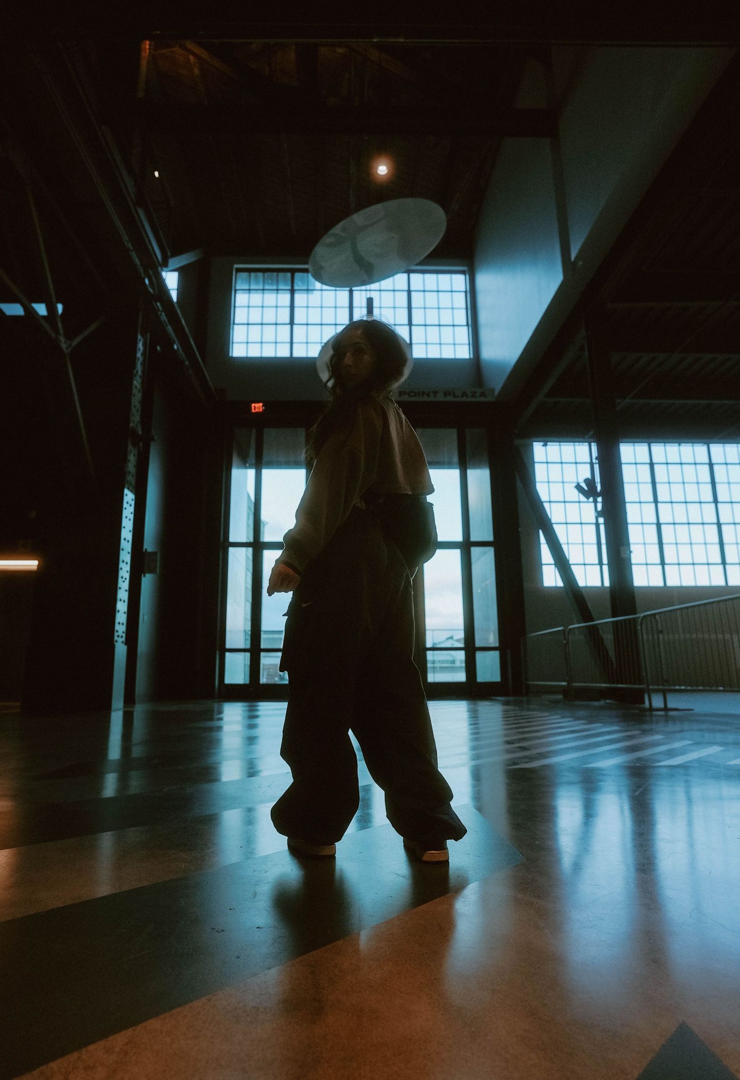 Person standing dramatically in dark industrial space with large windows and overhead light.