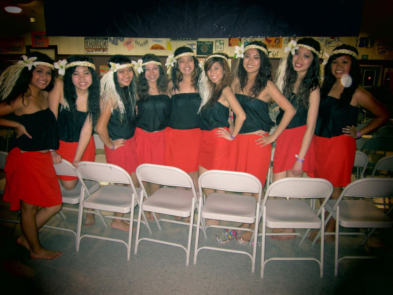 Group of women in matching black tops, red skirts, and floral headbands posing together at a Hawaiian hula dance performance.