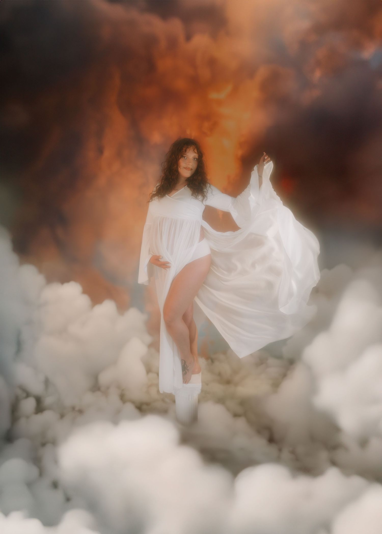 A pregnant woman in a flowing white dress stands majestically among dramatic clouds and fire sky.