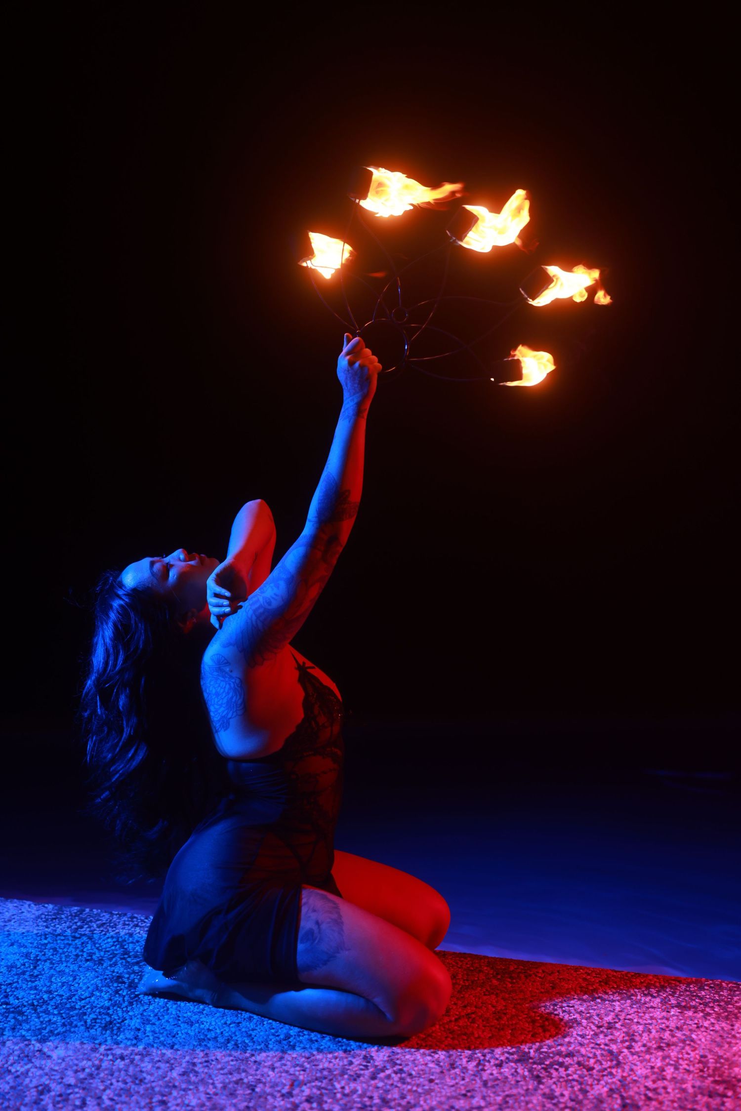 A woman kneels in blue and red light, holding a multi-flame fire prop aloft against a dark background.