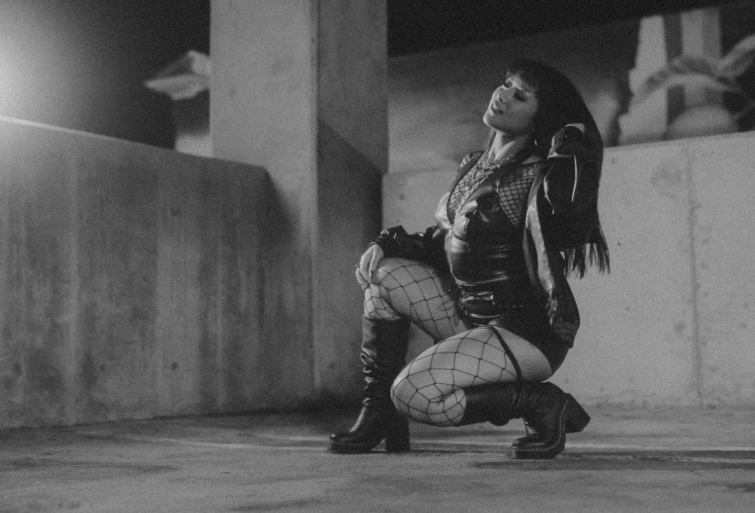 Black and white photo of woman in fishnet tights and leather jacket kneeling in parking garage.