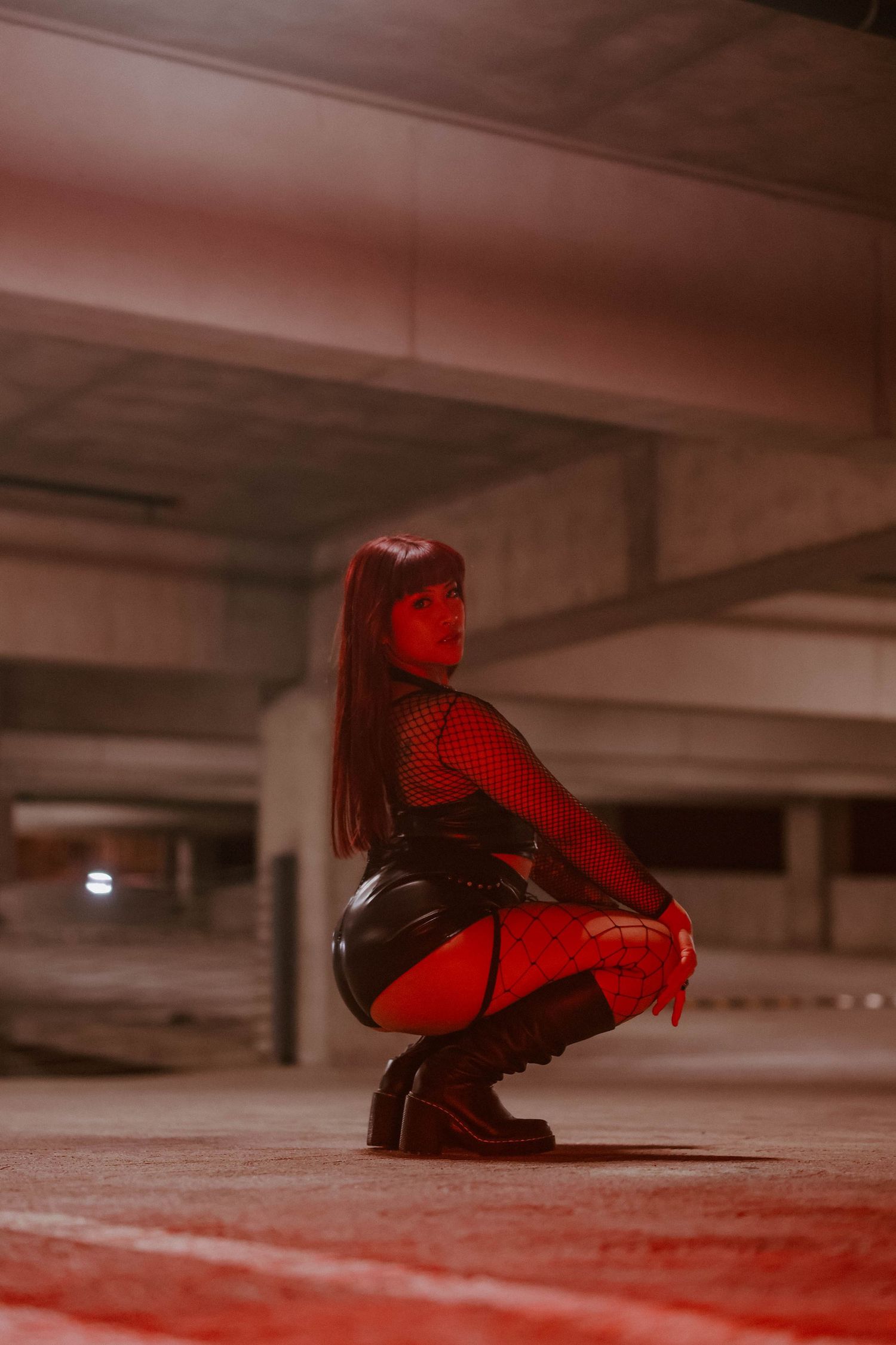 A woman in fishnet tights and leather skirt crouches in a red-lit parking garage.