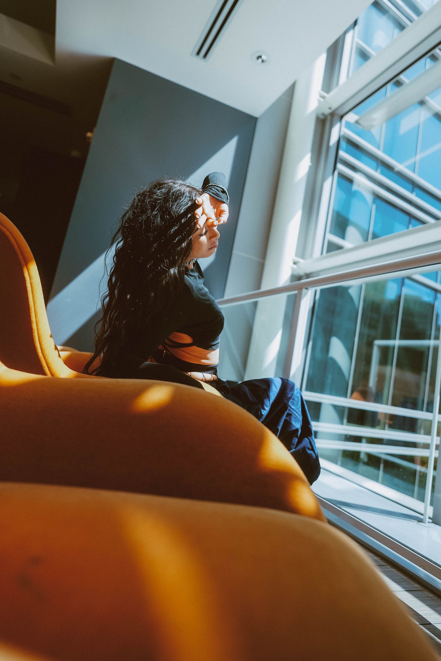Woman with long dark hair sits in a yellow chair gazing out large windows in a modern sunlit building interior.