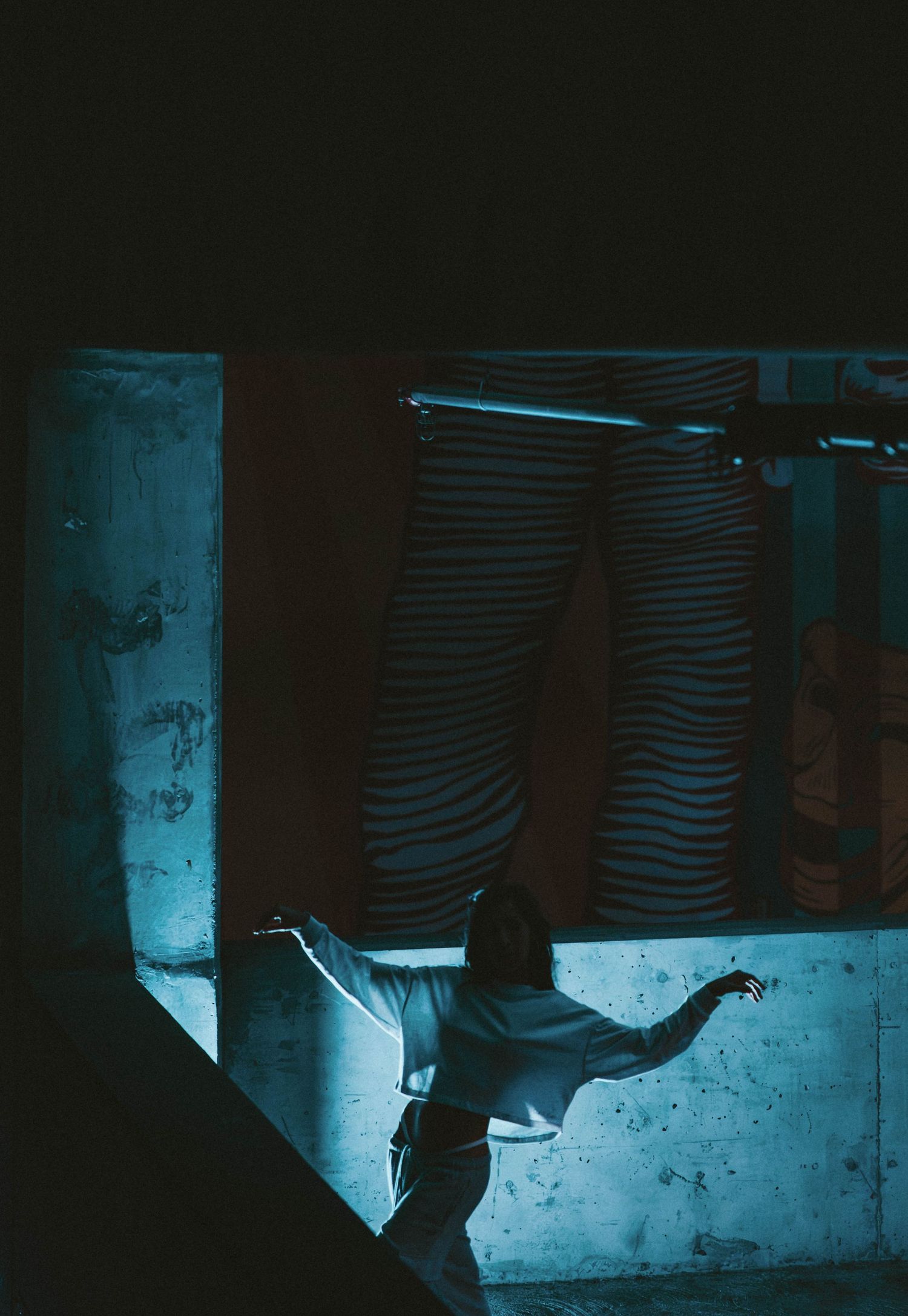 A dancer in white extends arms dramatically in a dark blue-lit industrial space.