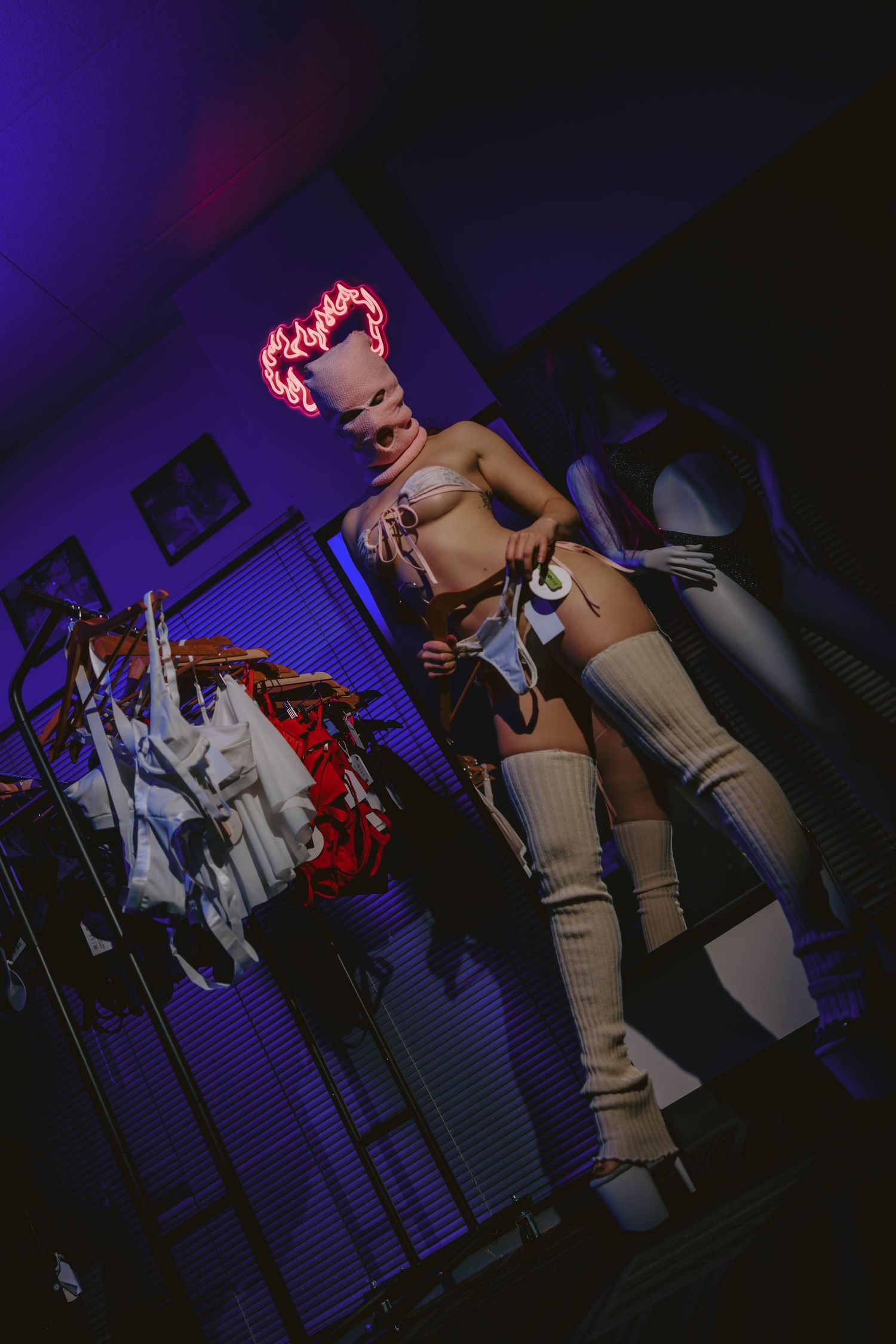 Masked performer in beige thigh-high boots and lingerie stands near clothing rack under neon light.