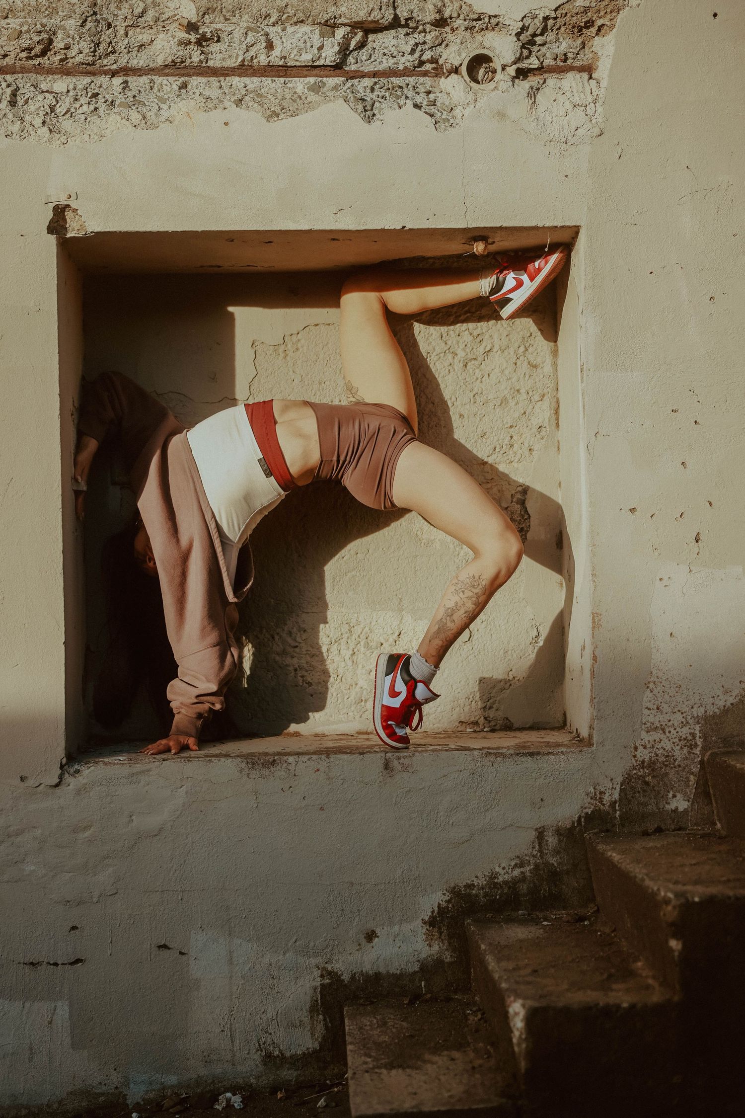 A flexible dancer poses dramatically in a rustic wall opening, wearing red Nike sneakers and brown shorts, with legs extended.