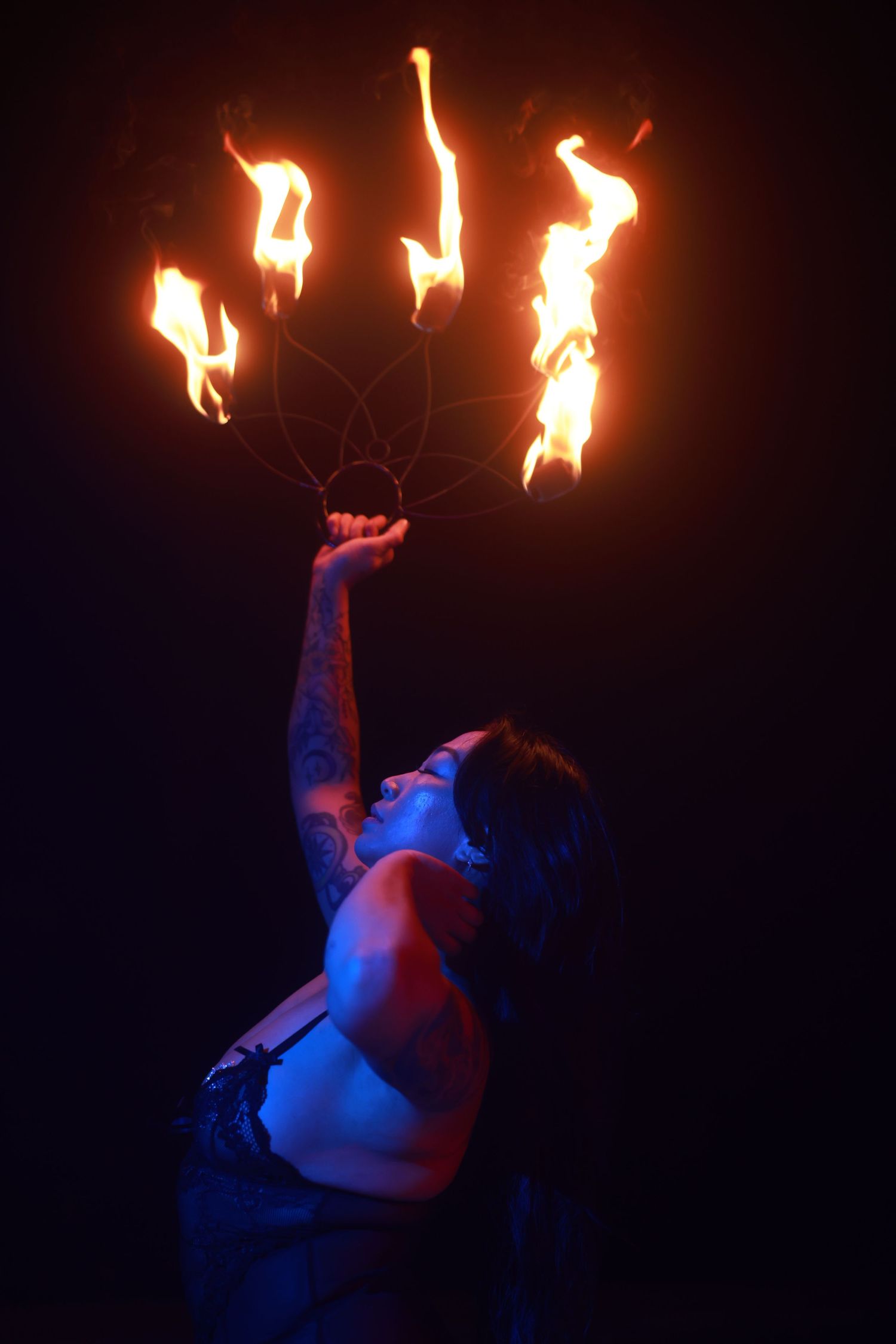 Tattooed fire performer holds flaming hoop aloft in dramatic dark stage setting with blue and red lighting.