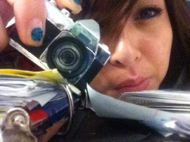 Woman with dark hair holds a small digital camera up to her eye, with blue nail polish visible on her fingers.