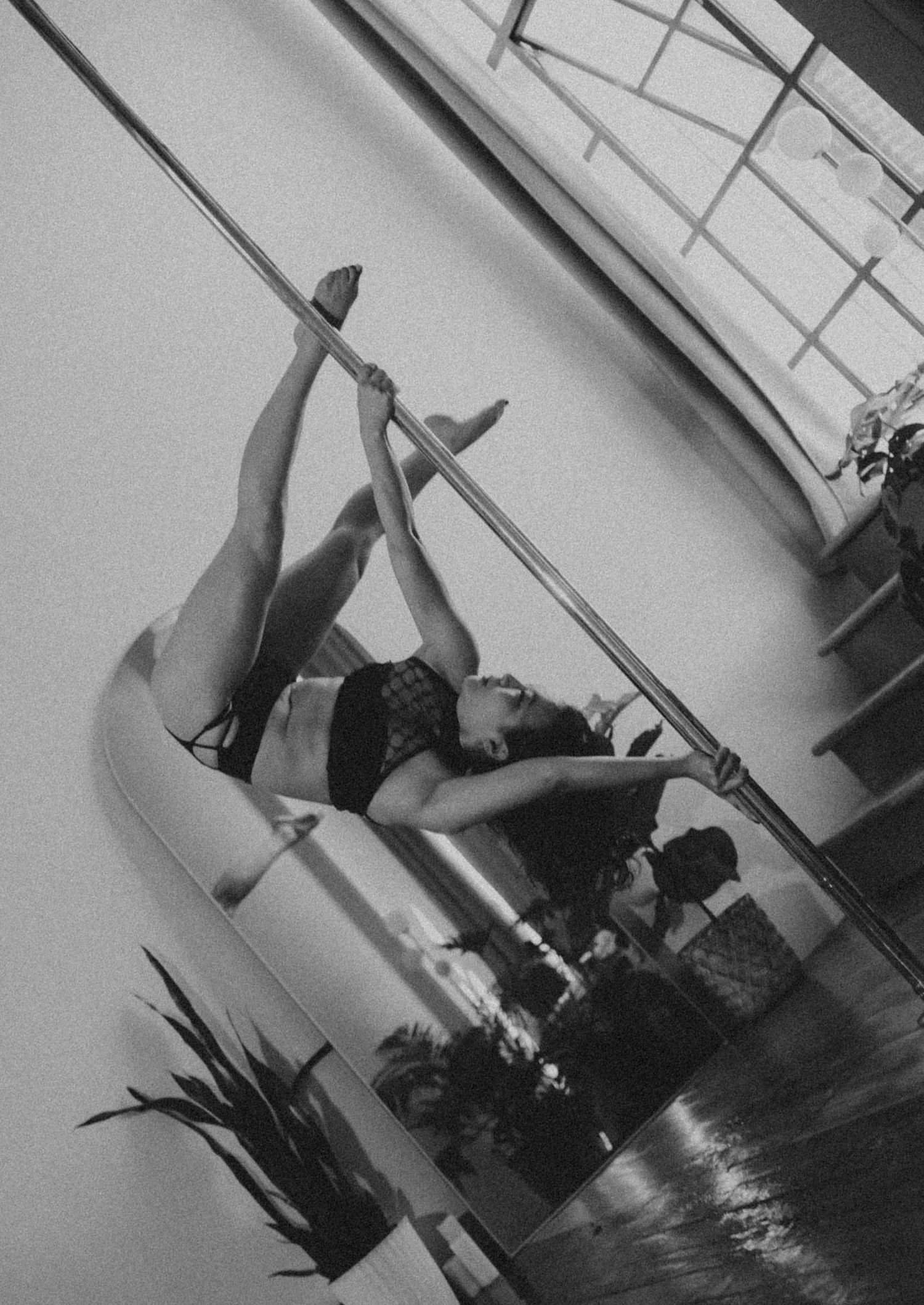 Black and white photo of a woman performing an advanced pole dance move in a studio.