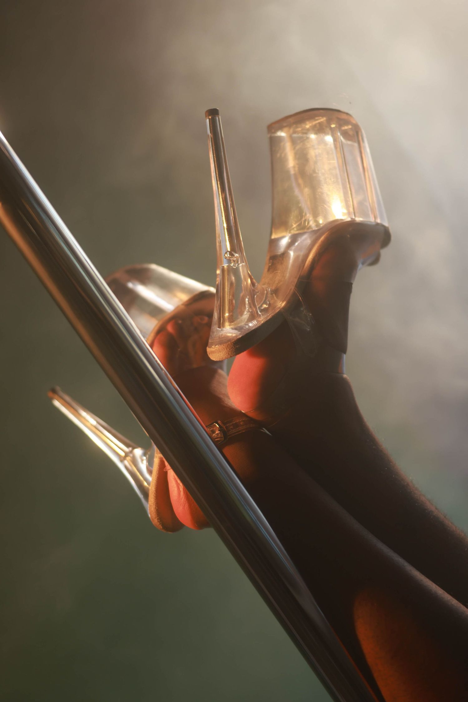 Close-up of clear platform high heels gripping a shiny pole under warm amber stage lighting.