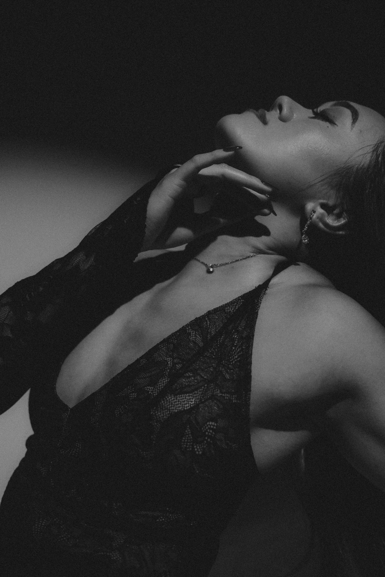 Black and white photo of a woman in lace bodysuit and gloves, posing gracefully with hand near neck, wearing a delicate necklace.