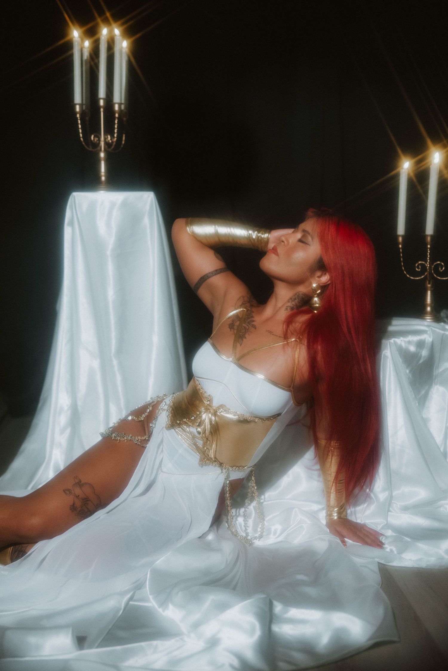 Red-haired woman in white and gold goddess costume reclines on white satin with candelabras.
