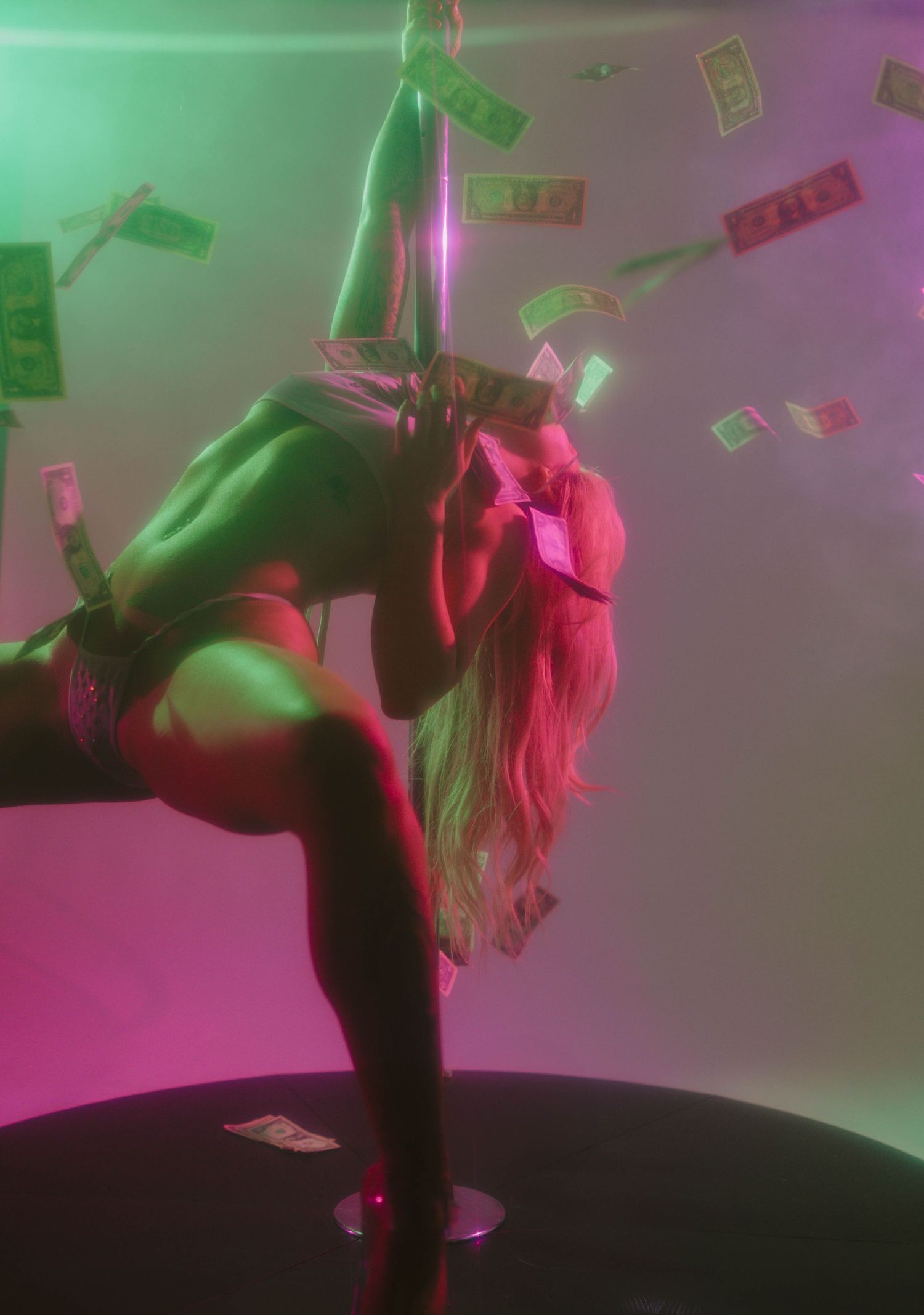 Pole dancer performing with money raining down under pink and green neon lights in a nightclub.