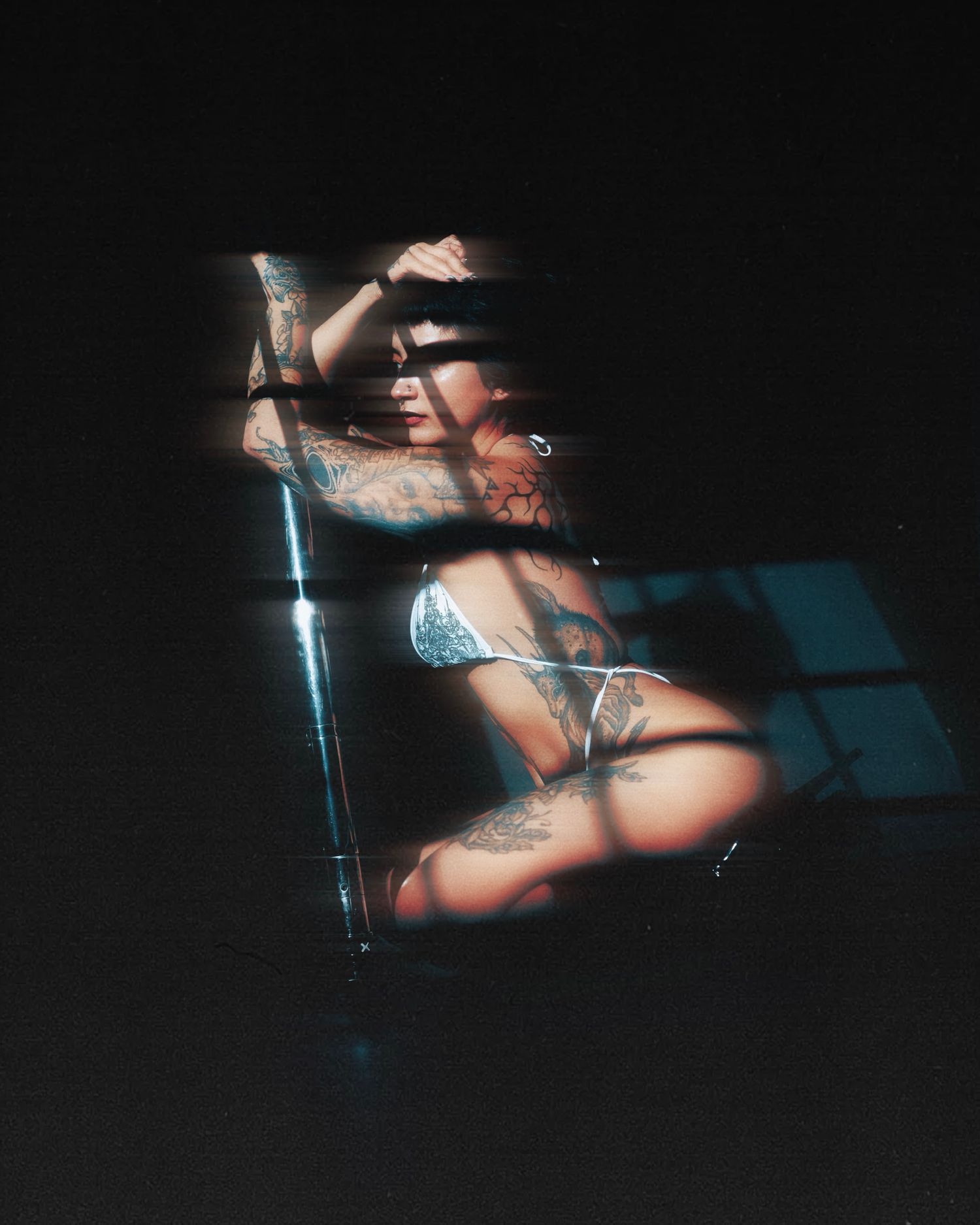 Tattooed woman posing in lingerie with dramatic shadow light patterns creating artistic contrast.