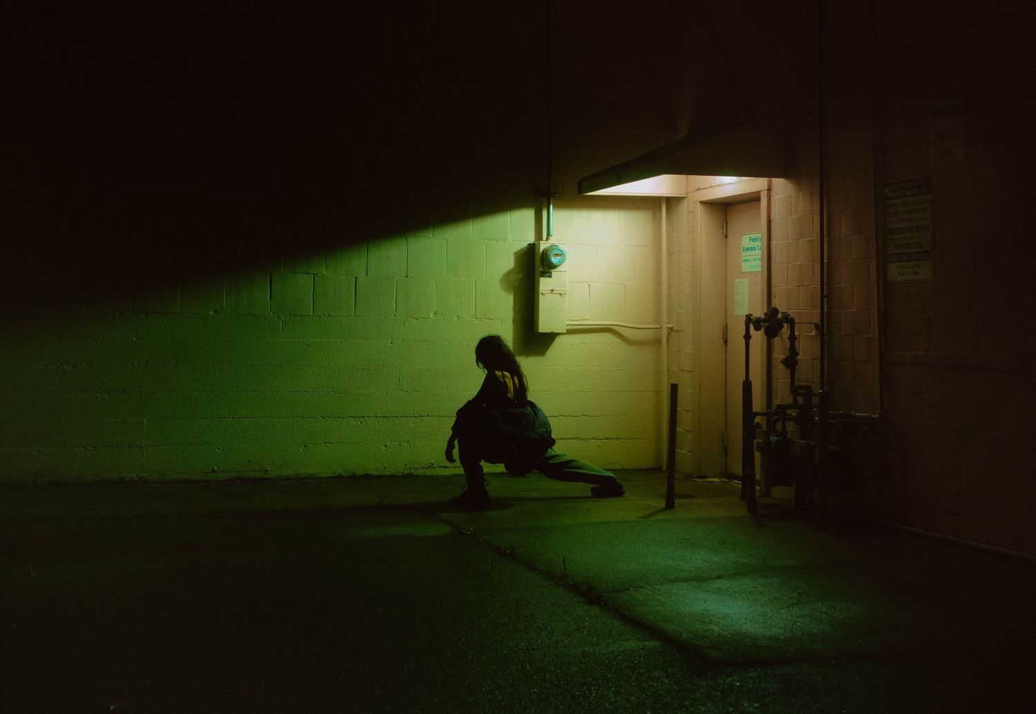 Person crouching alone under green neon light in dark urban alley at night.
