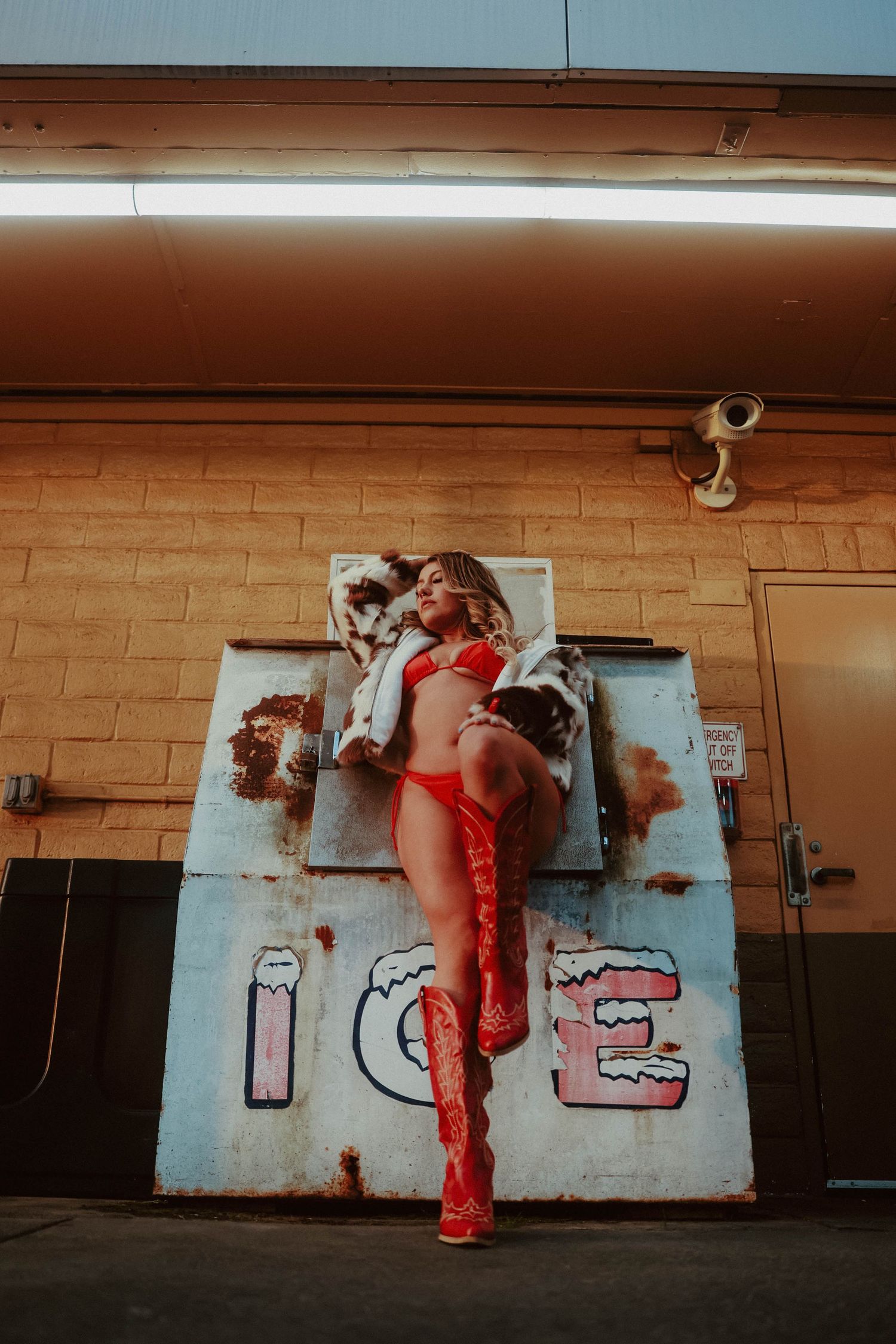 Woman in red lingerie and boots poses against ICE machine under neon light.