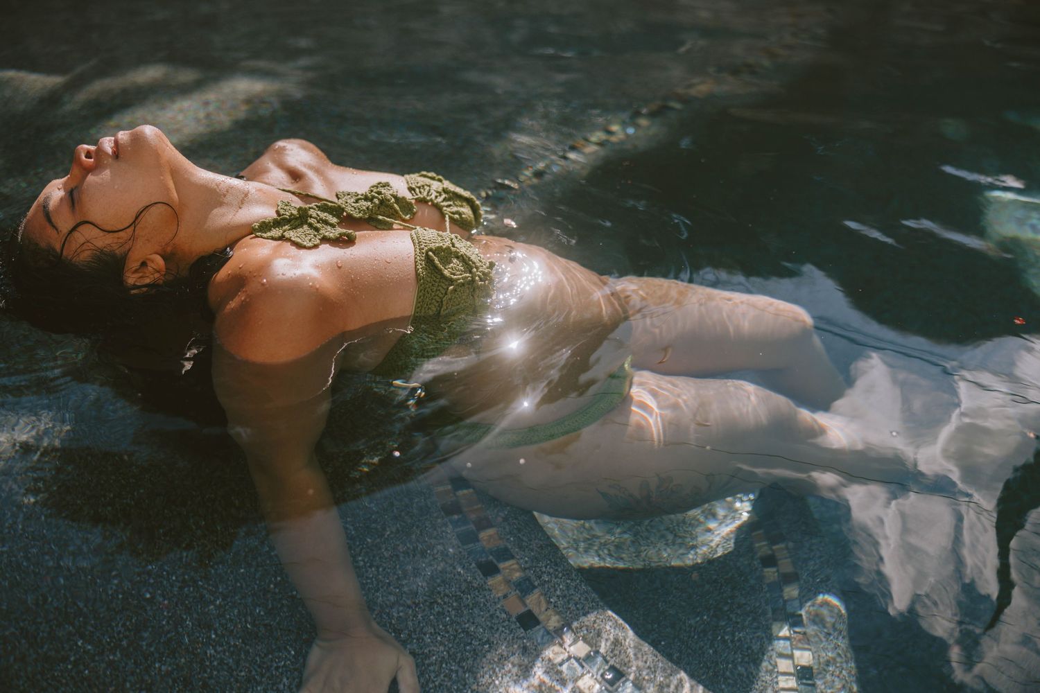 Woman in green bikini top floating gracefully in a sunlit pool, eyes closed, wearing a sheer white skirt.