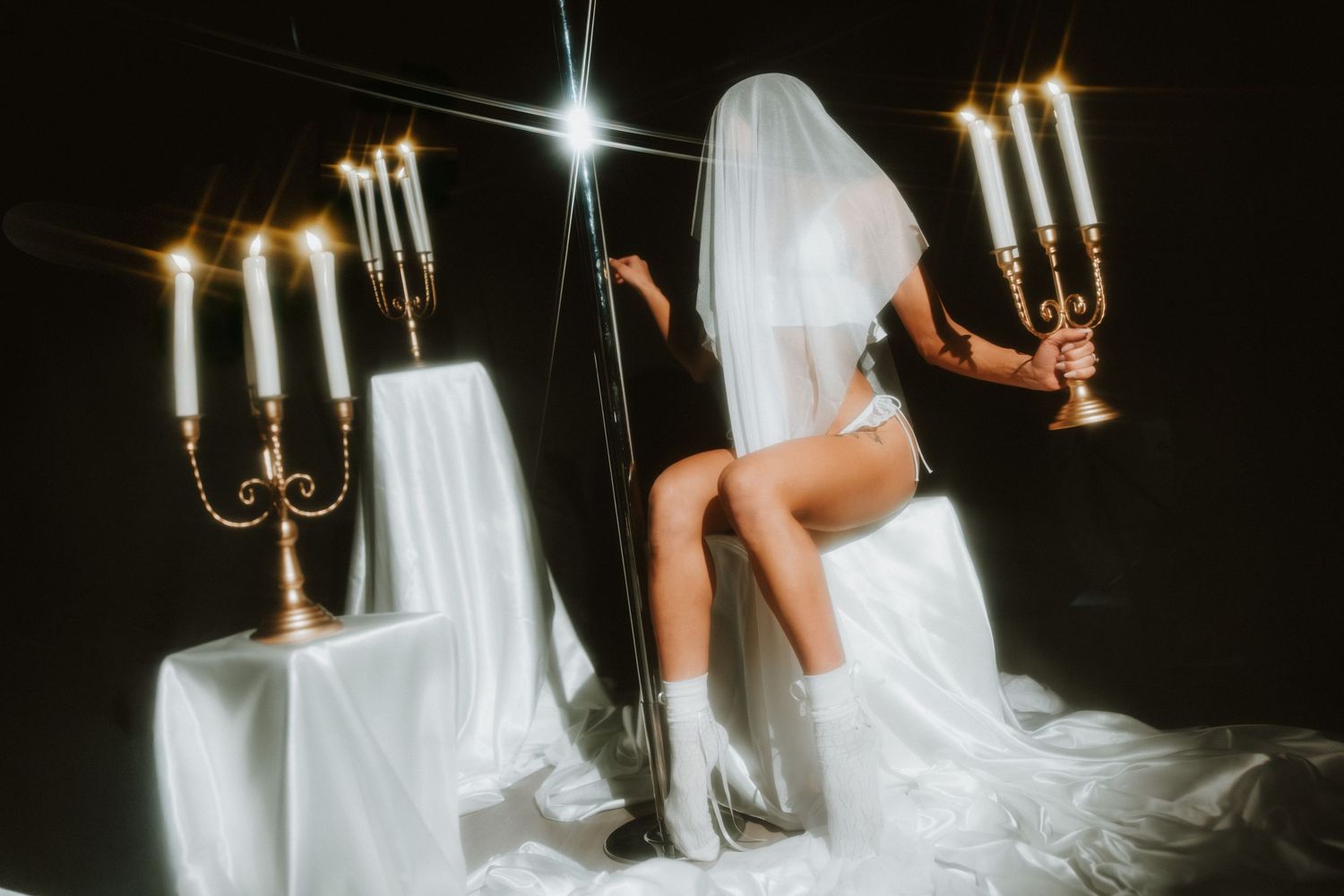 Bride in white veil and lingerie sits between golden candelabras with lit candles on draped tables in dark studio.