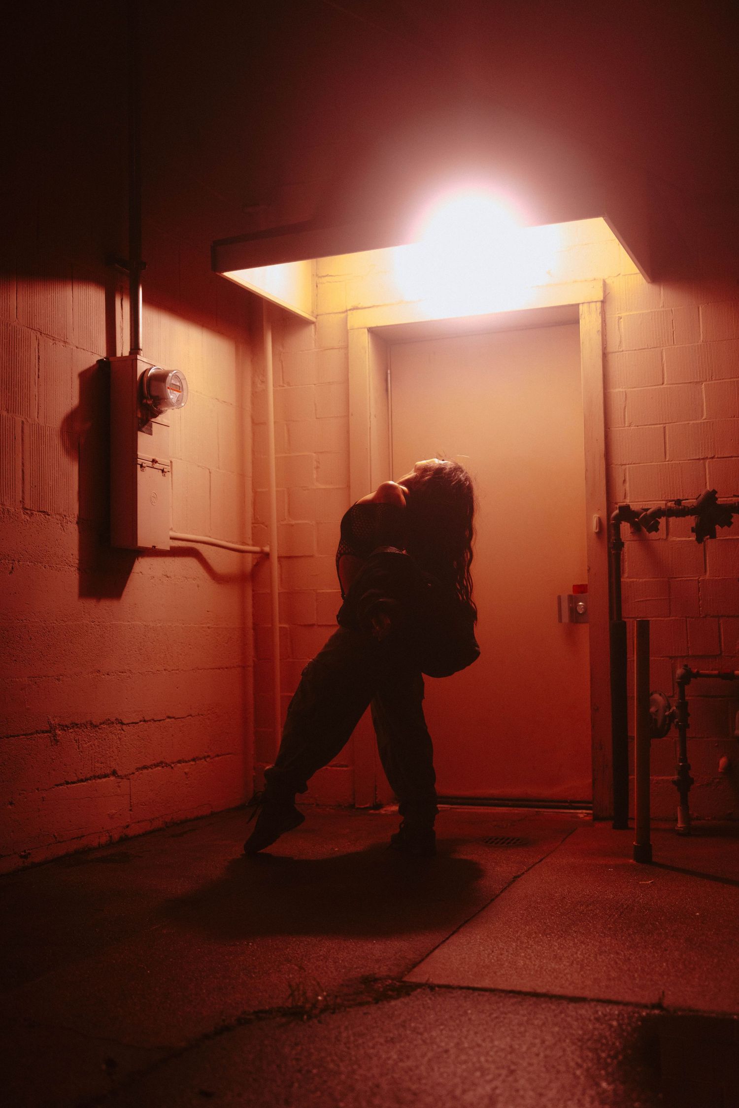 Silhouette of a person dancing under a glowing light by a door in a dark, red-lit alley at night.