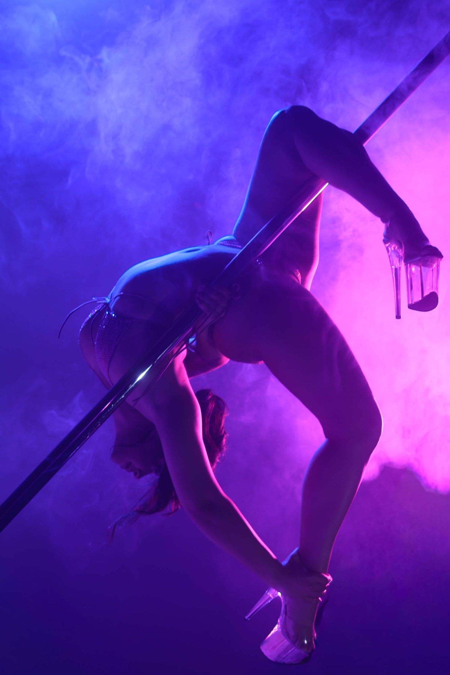 Pole dancer performing acrobatic move in purple smoky neon-lit studio environment.