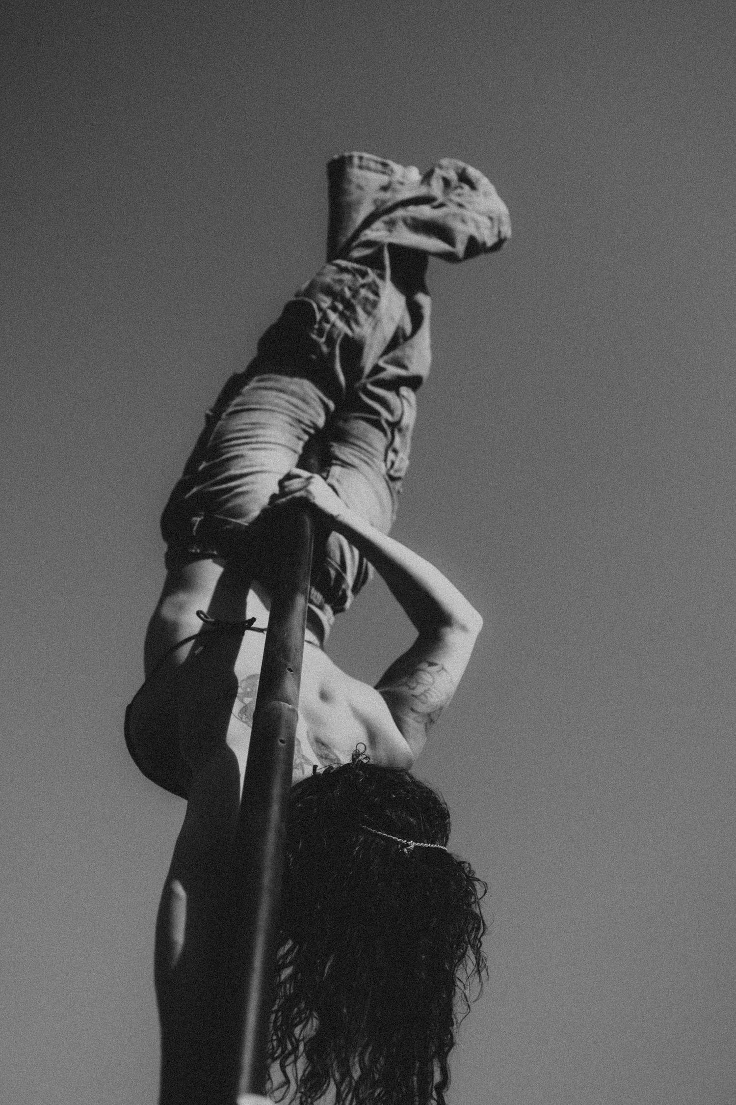 Black and white photo of a shirtless person performing an acrobatic pole move against a clear sky.
