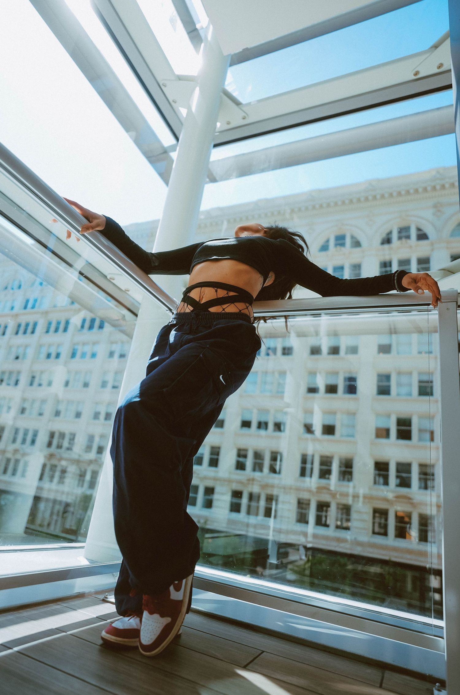 Woman in crop top and wide-leg pants posing by large windows with urban building view.