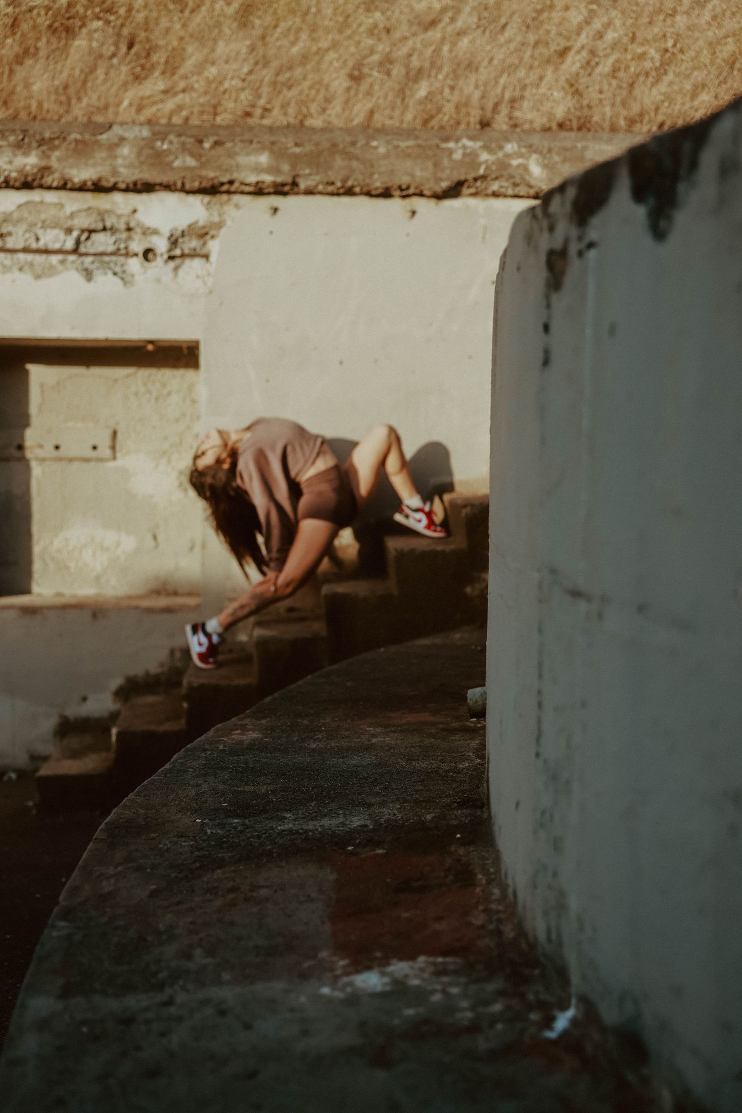 Woman in brown outfit with red sneakers crouching on curved stone steps between concrete walls.