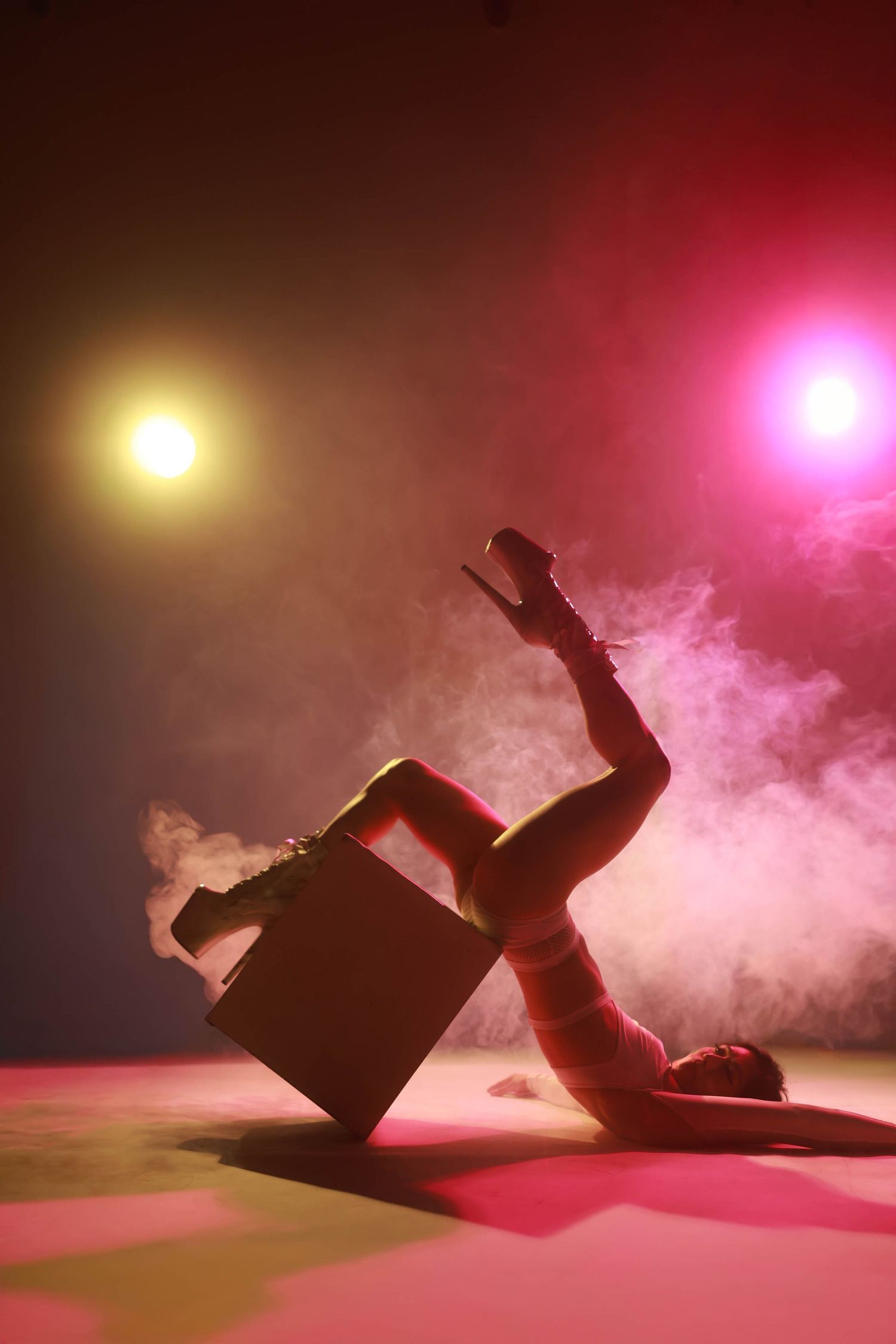 A female dancer poses dramatically on stage floor with a prop box under colorful smoky stage lights.