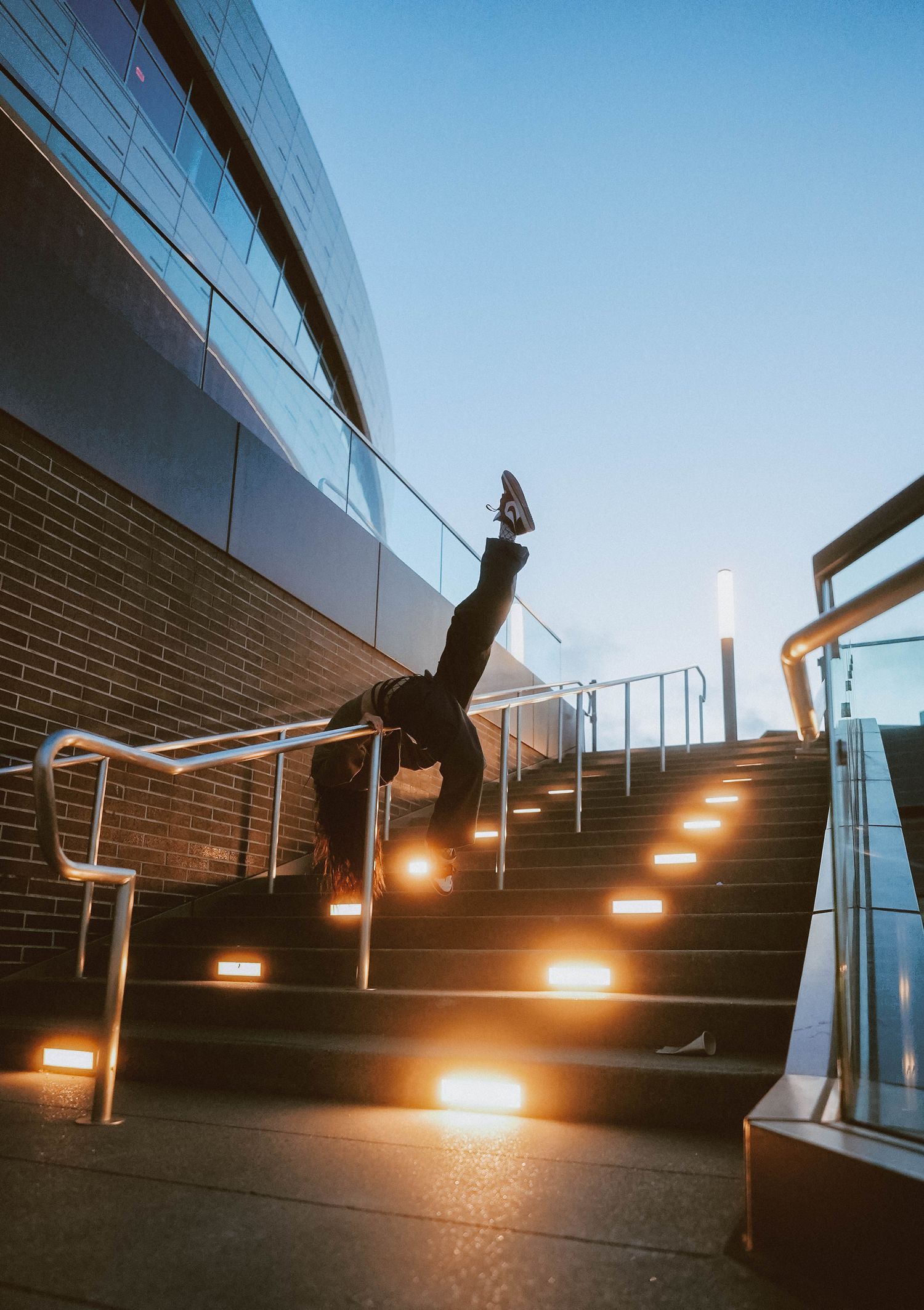 Person performing a handstand on illuminated outdoor stairs at dusk near a modern glass building.