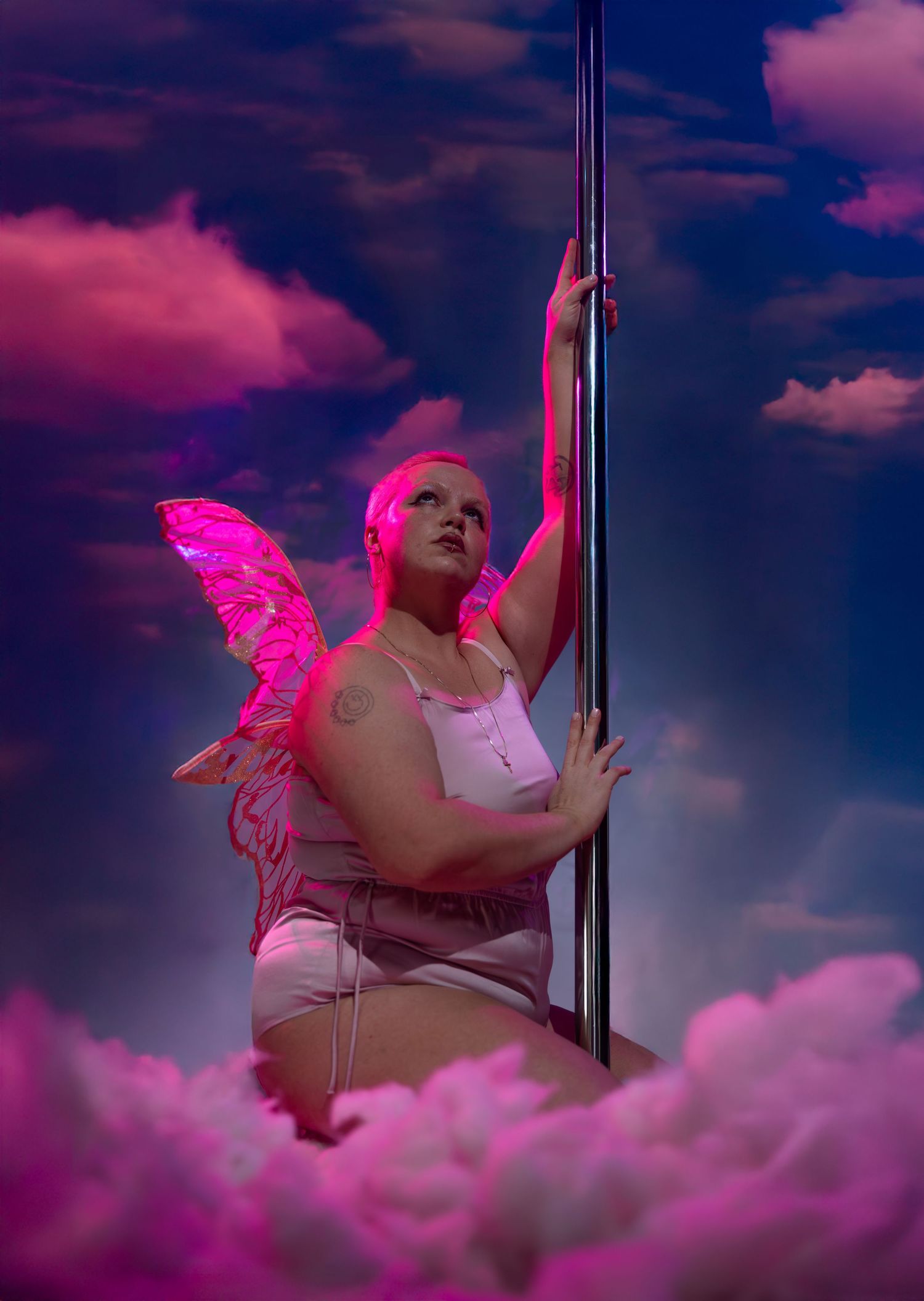 Plus-size woman with pink fairy wings poses on pole against dreamy pink cloudy sky backdrop.