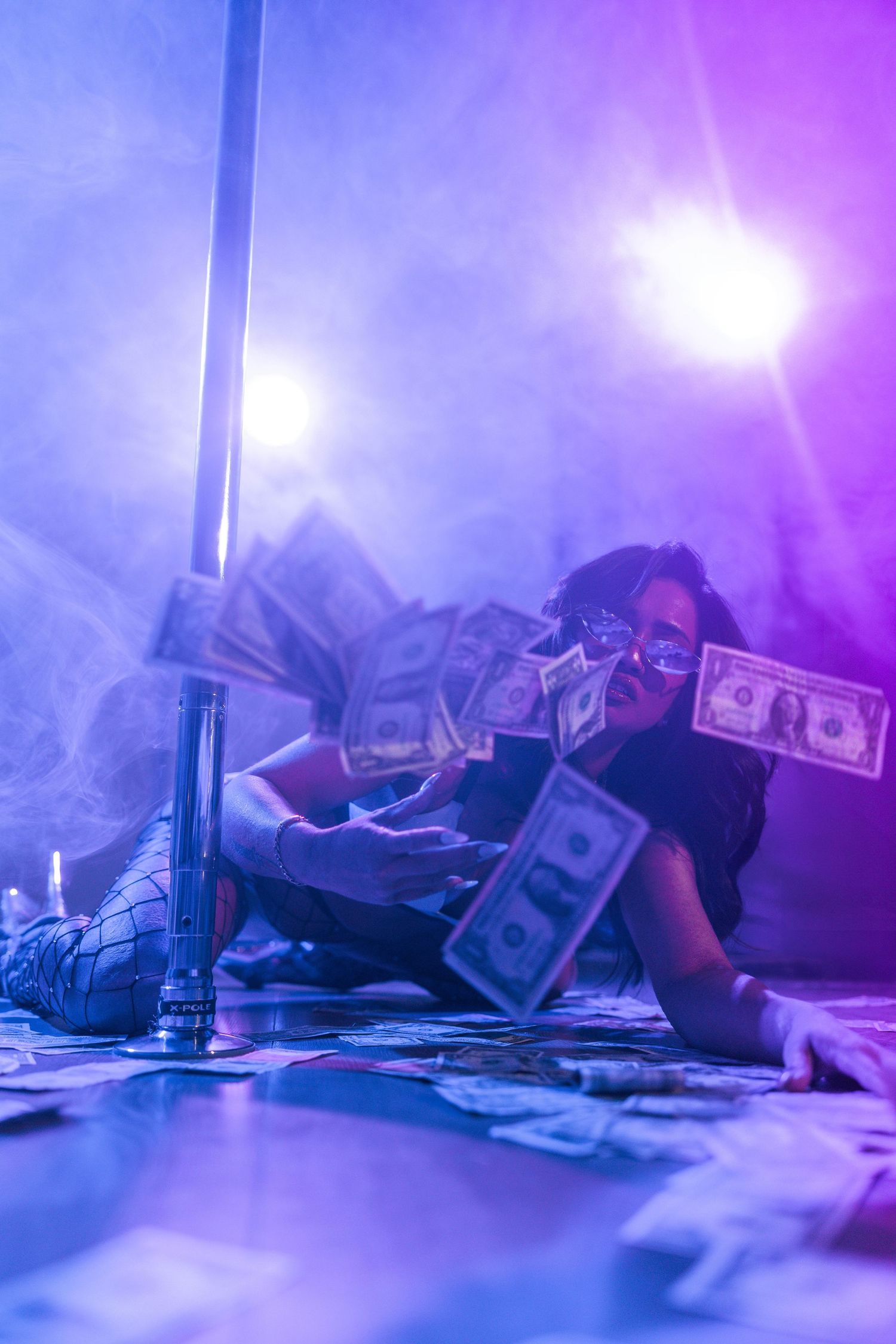 A woman poses on a smoke-filled stage floor covered in dollar bills under purple neon lights.