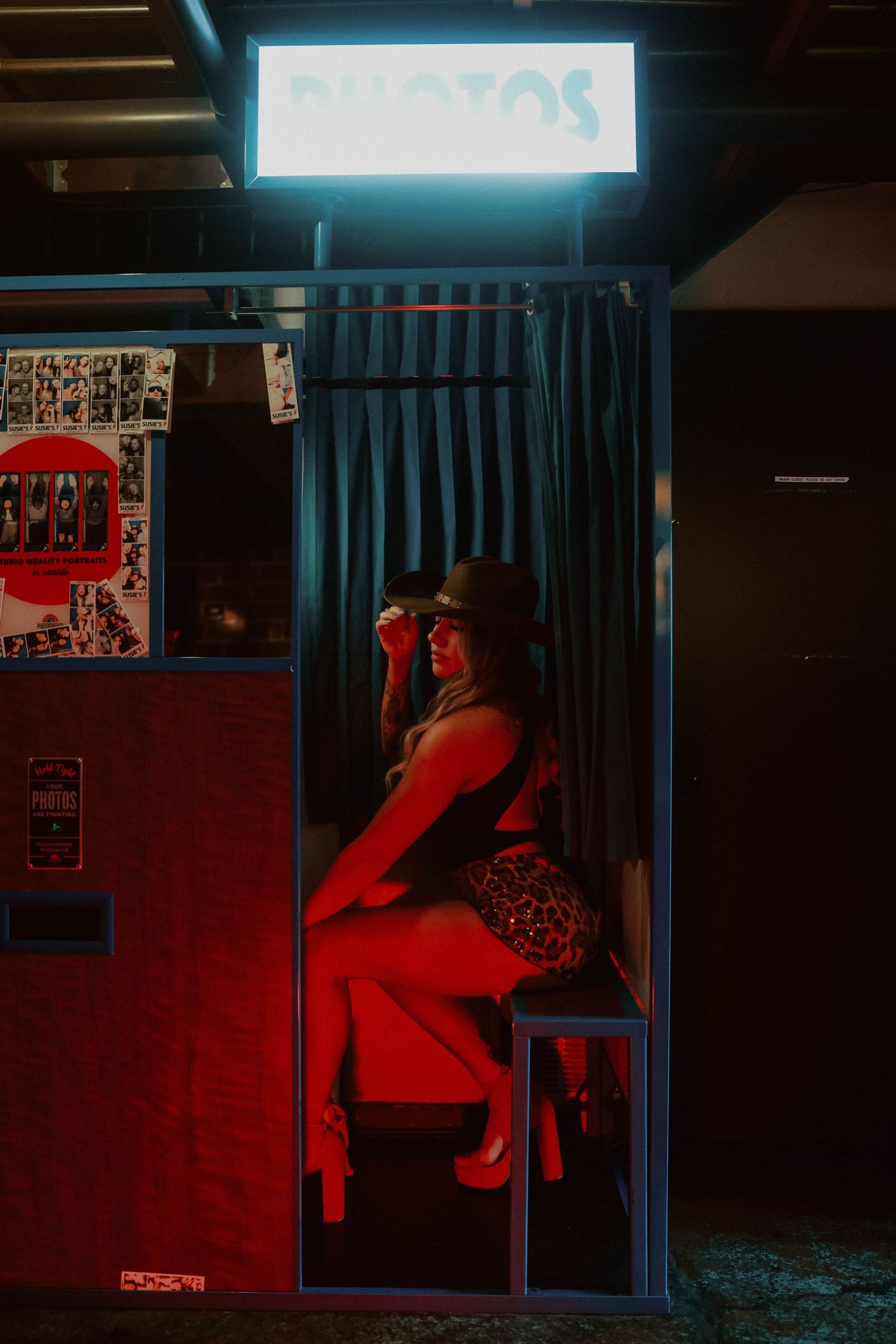 Woman in leopard skirt and cowboy hat sitting inside a glowing photo booth at night under neon sign.