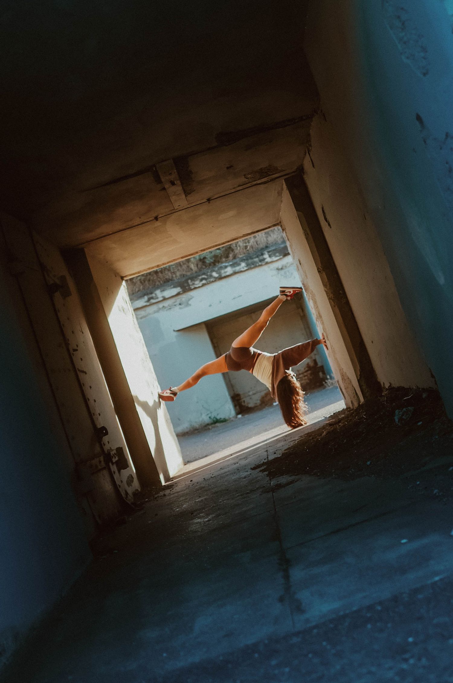 Dancer performs a dramatic split pose in a dark tunnel doorway of an abandoned building with natural light.