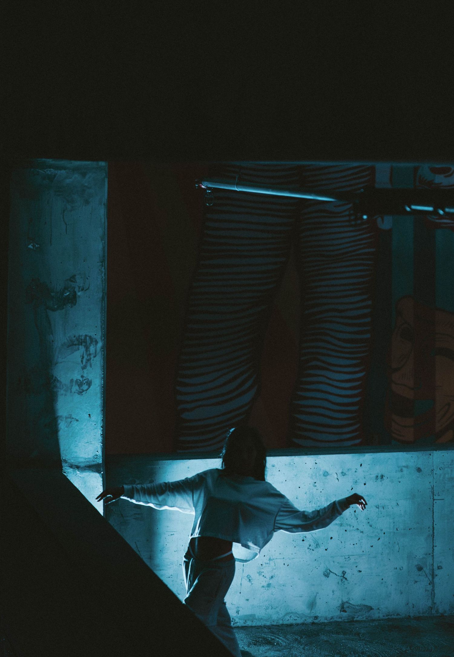 A dancer poses dramatically in a dark industrial space with blue lighting and striped fabric backdrop.
