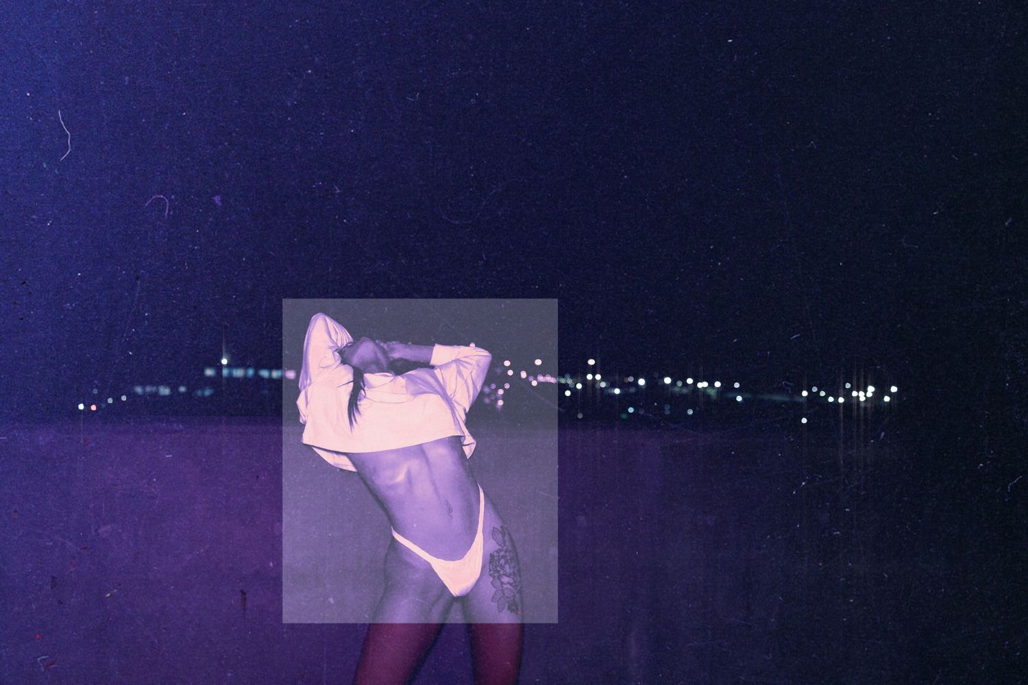 Person posing outdoors at night with city lights in the background under a purple-toned atmosphere.