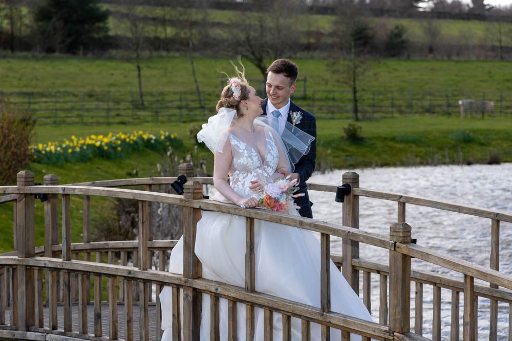 Bride and groom share a romantic moment on a wooden bridge overlooking a pond, surrounded by spring daffodils.