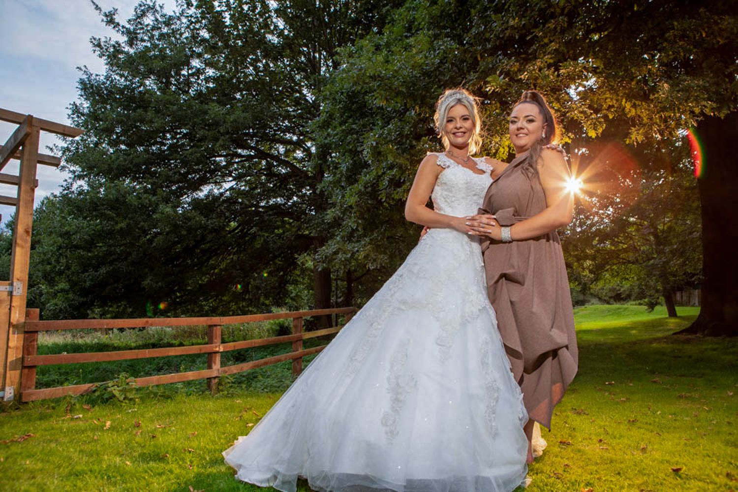 Bride in white gown poses with bridesmaid in taupe dress outdoors near wooden fence with sunburst through trees.