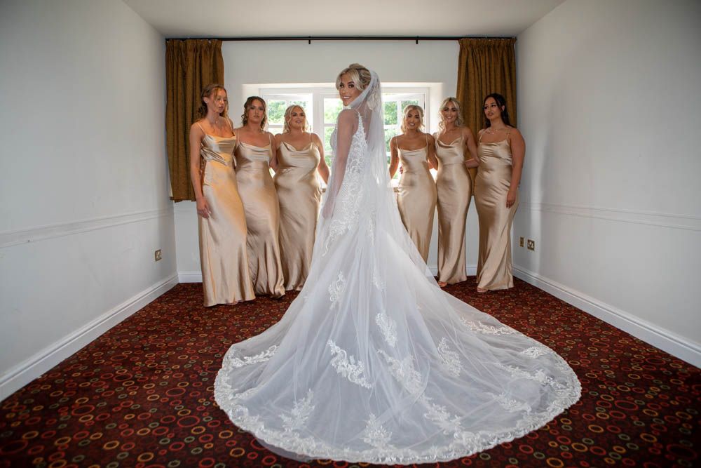Bride in lace cathedral veil stands before five bridesmaids in gold satin gowns indoors.