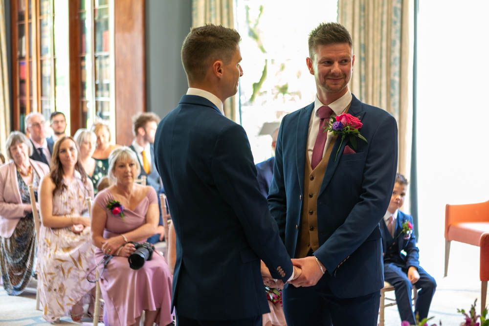 Two grooms holding hands at wedding ceremony with guests seated in background.