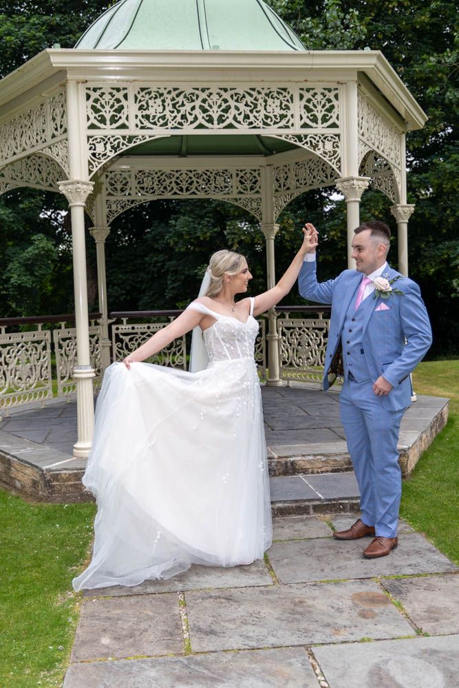 Bride and groom dancing by ornate white gazebo outdoors on their wedding day, bride in white gown, groom in blue suit.