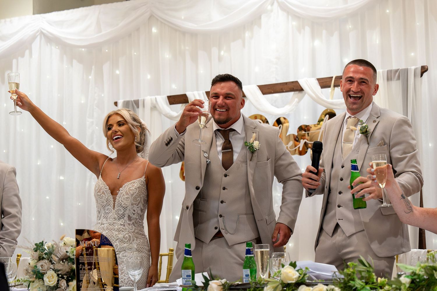 Bride and groom celebrate with best man at wedding reception, raising champagne glasses and smiling joyfully.