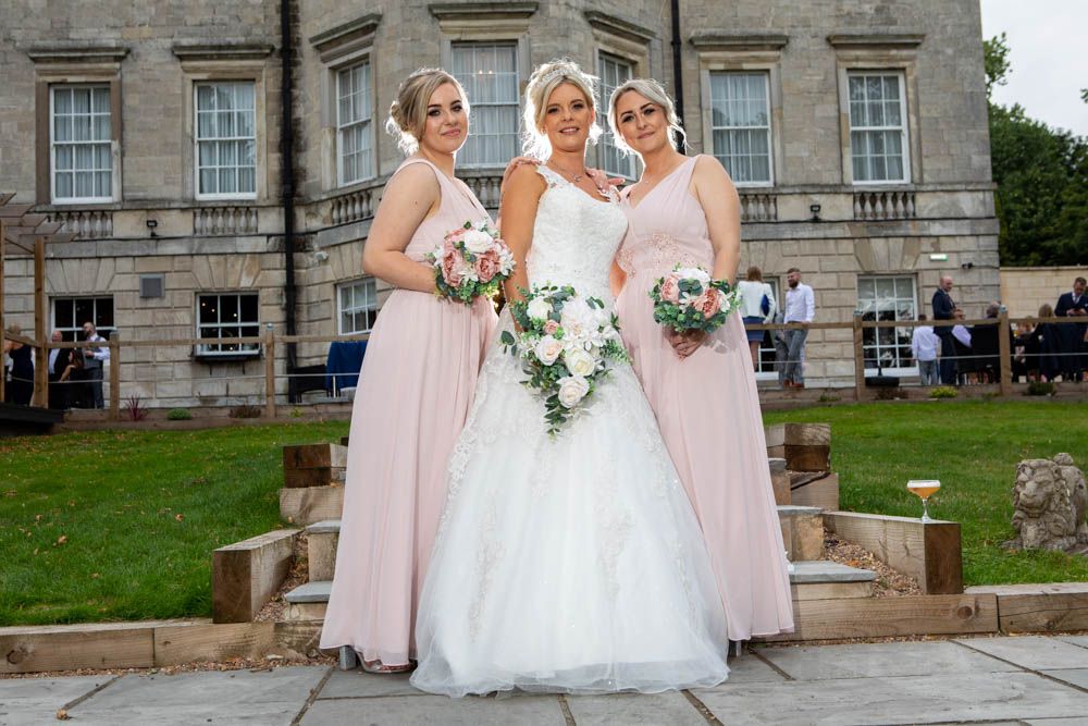 Bride in white gown with two bridesmaids in blush pink dresses holding bouquets outside a grand stone manor - Aston Hall Hotel.