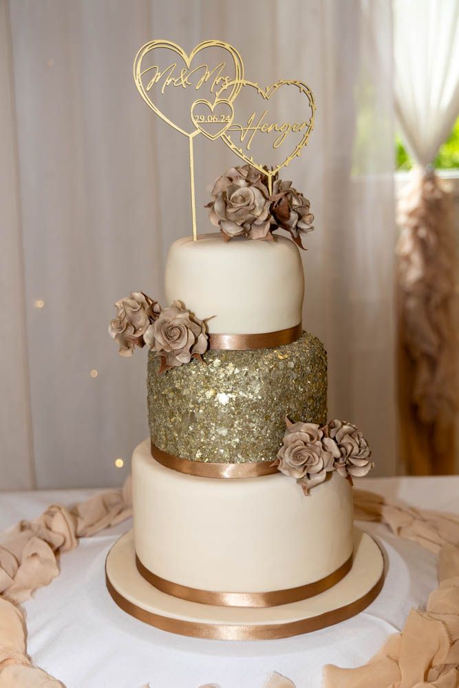 Elegant three-tier wedding cake with gold sequin middle tier, mauve roses, and gold Mr & Mrs heart cake topper.