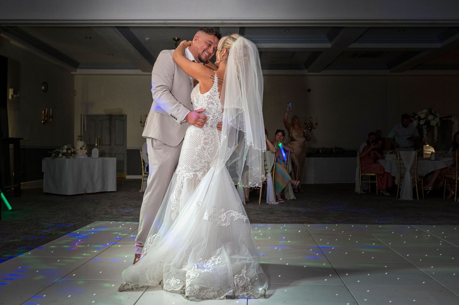 Bride and groom share romantic first dance on glittering dance floor, bride in lace gown with veil, groom in gray suit.