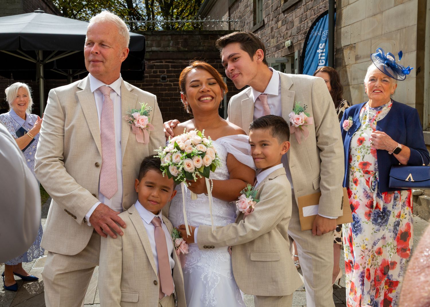 Wedding party poses outdoors, bride in white gown holds bouquet, flanked by men in beige suits and two young boys.