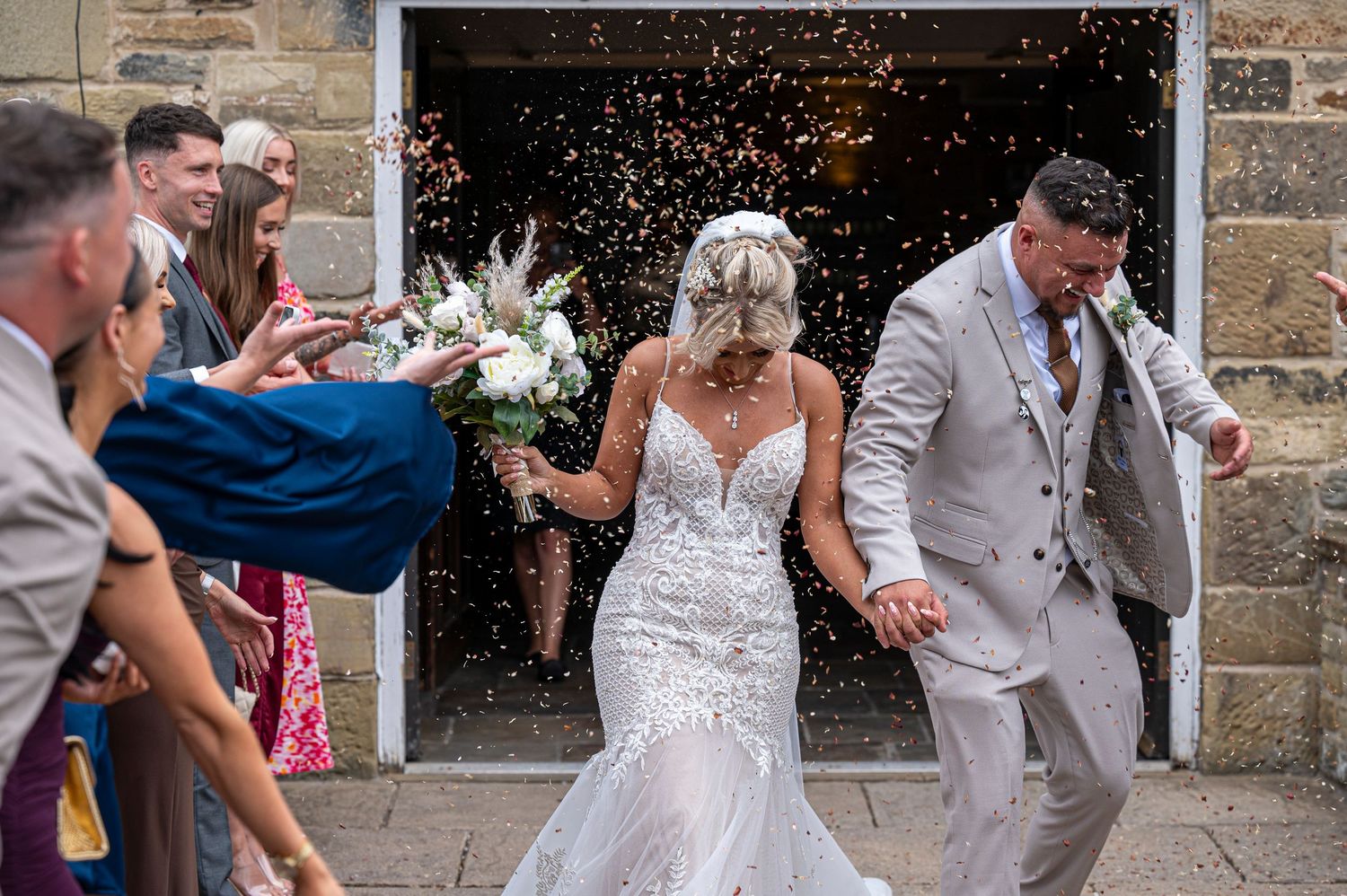 Bride and groom exit church ceremony through confetti shower, bride holding floral bouquet in lace wedding dress.